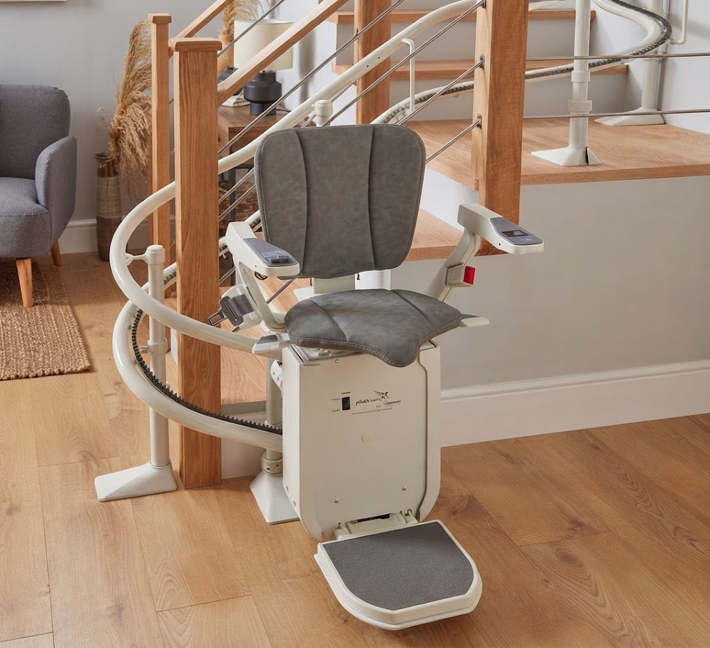 Ein moderner Treppenlift mit anthrazitfarbenem Sitz parkt platzsparend an einer weißen Führungsschiene im unteren Bereich einer Holztreppe. Das elegante Design fügt sich harmonisch in den hellen, lichtdurchfluteten Wohnraum ein.